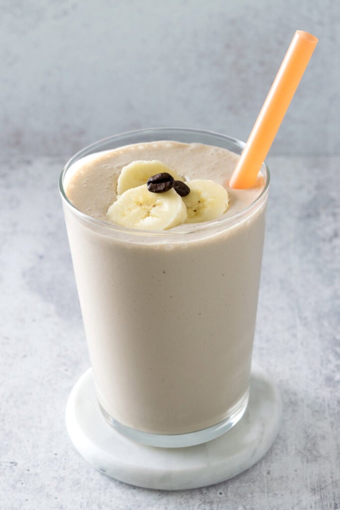 Creamy coffee banana smoothie in a clear glass that's sitting on a coaster. The smoothie is garnished with three slices of banana and a coffee bean and has an orange straw in it.