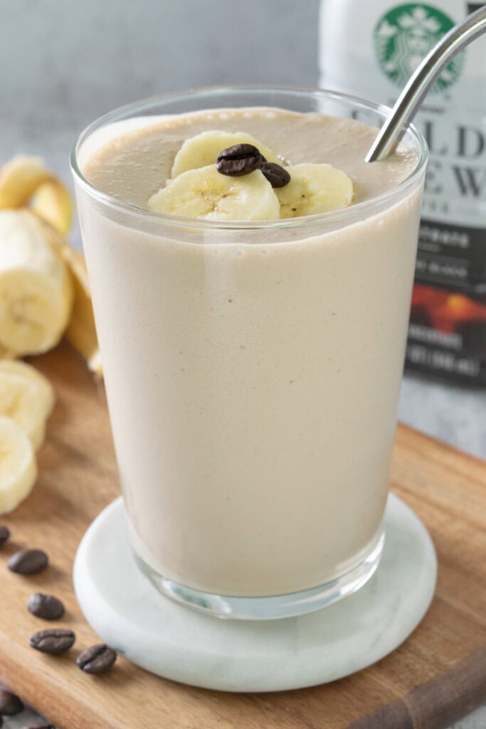A coffee banana smoothie topped with banana slices and coffee beans in a glass with straw. The smoothie is sitting next to a sliced banana and bottle of cold brew coffee.