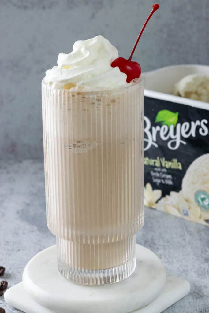 Coffee milkshake garnished with whipped cream and a cherry next to a carton of vanilla ice cream.