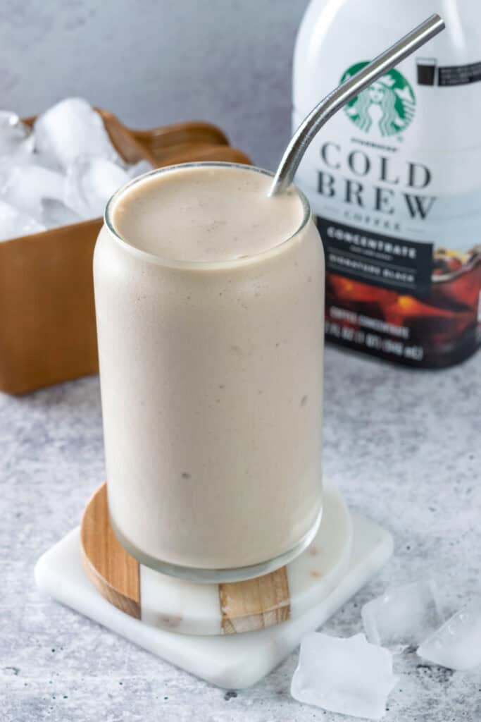 A frozen coffee drink in a glass with straw, next to a bottle of cold brew concentrate and a scoop of ice cubes.