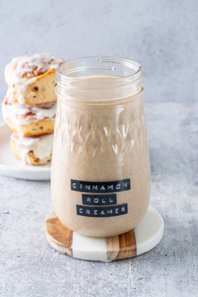 Jar of homemade cinnamon roll coffee creamer next to a stack of three cinnamon rolls dripping with glaze.