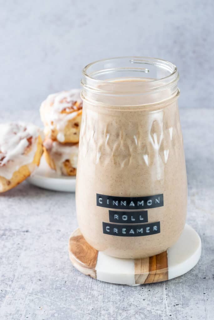 Homemade cinnamon roll coffee creamer in a fluted mason jar with a black embossed label on it. The jar of creamer is on a brown and white coaster and is in front of a plate of homemade cinnamon buns.