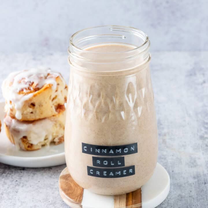 Jar of homemade cinnamon roll coffee creamer next to two cinnamon buns dripping with glaze.