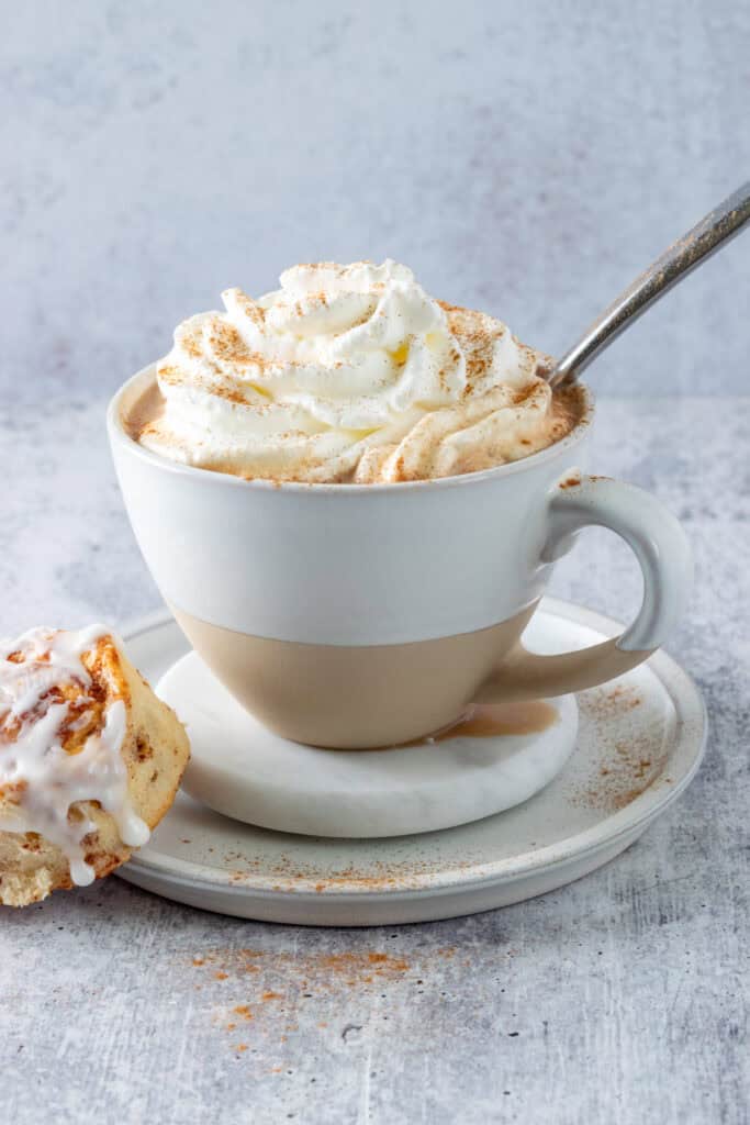 A homemade cinnamon roll latte topped with whipped cream and cinnamon with a cinnamon bun leaning on the plate next to the coffee cup.