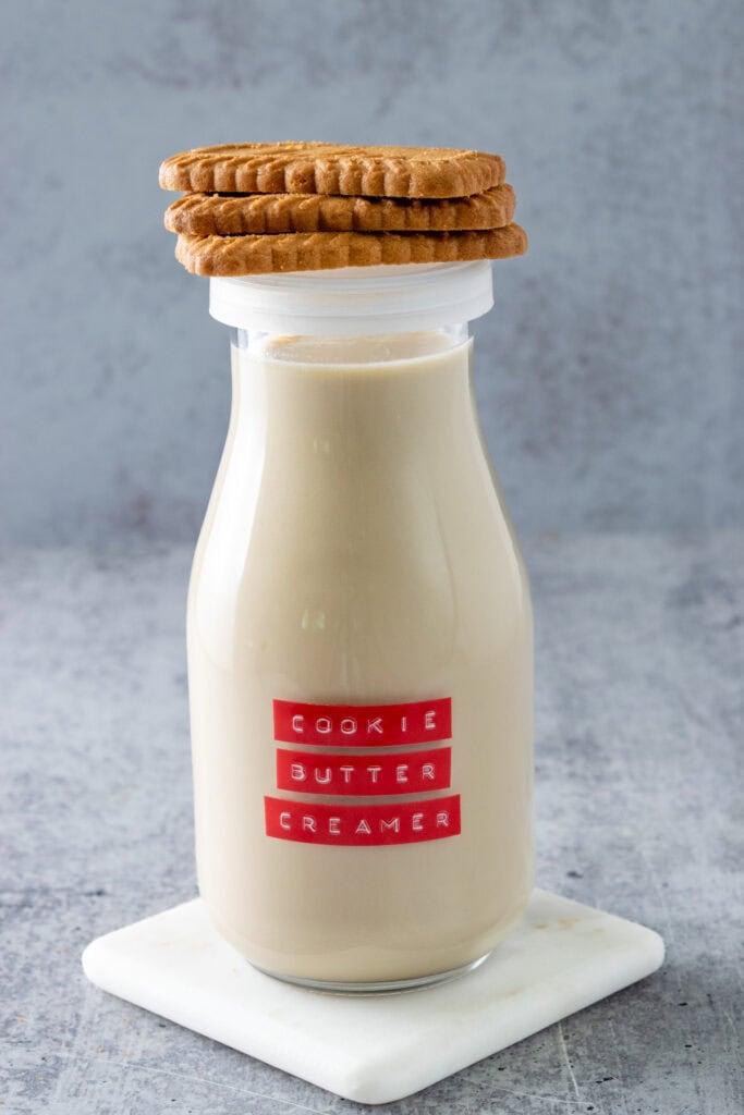 Small milk bottle with red cookie butter creamer label on it, that's filled with the homemade coffee creamer and has 3 Biscoff cookies stacked on top of the cap.