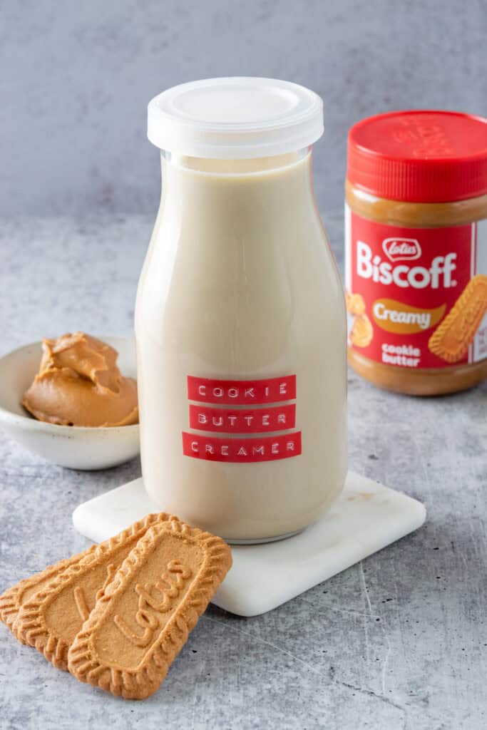 Three Lotus Biscoff speculoos cookies, a jar of Biscoff creamy cookie butter and a small bowl containing a dollop of cookie butter, all next to a bottle of homemade cookie butter coffee creamer.