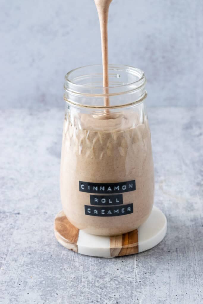 Homemade cinnamon roll coffee creamer being poured into a glass mason jar with a label on it that says, cinnamon roll creamer.