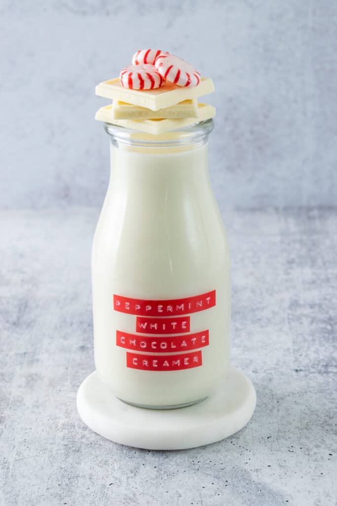 A full bottle of homemade peppermint white chocolate coffee creamer, that has a red label on it that says, peppermint white chocolate creamer, and has three white chocolate squares and three starlight peppermint candies stacked on top of it.