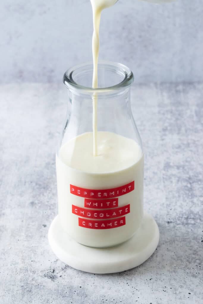 Stream of white chocolate coffee creamer being poured into a small milk bottle that has a red label on it that says, peppermint white chocolate creamer.