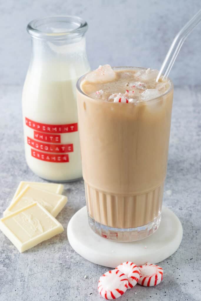 An iced peppermint mocha with pieces of crushed peppermint candy on top, sitting next to three starlight peppermint candies, three white chocolate Ghirardelli squares and a bottle of homemade peppermint white chocolate creamer.