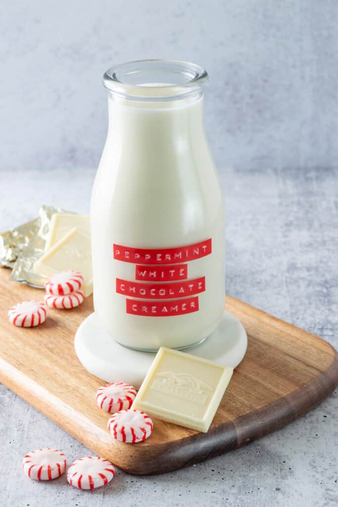 A bottle of homemade peppermint white chocolate creamer with a red and white embossed label on it. The bottle of creamer is sitting on a white marble coaster which is on a wood cutting board surrounded by Ghirardelli white chocolate squares and peppermint candies.