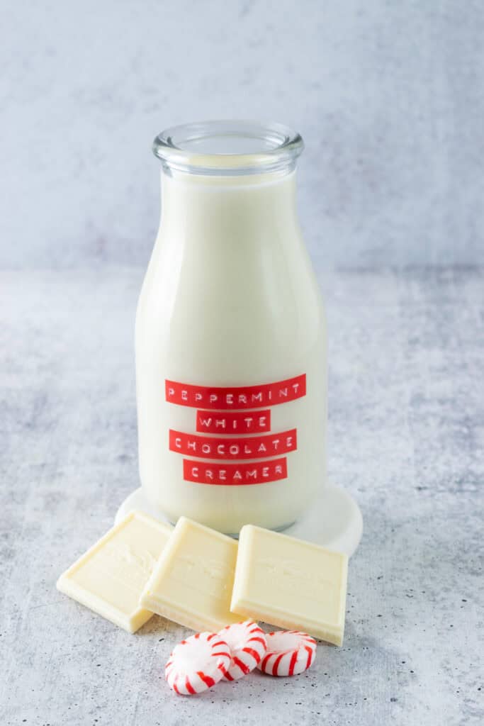 Bottle of homemade peppermint white chocolate coffee creamer with a red label on it, next two three squares of white chocolate and a few peppermint candies.