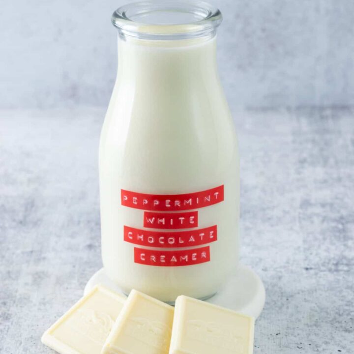 Bottle of homemade peppermint white chocolate coffee creamer with a red label on it, next two three squares of white chocolate and a few peppermint candies.