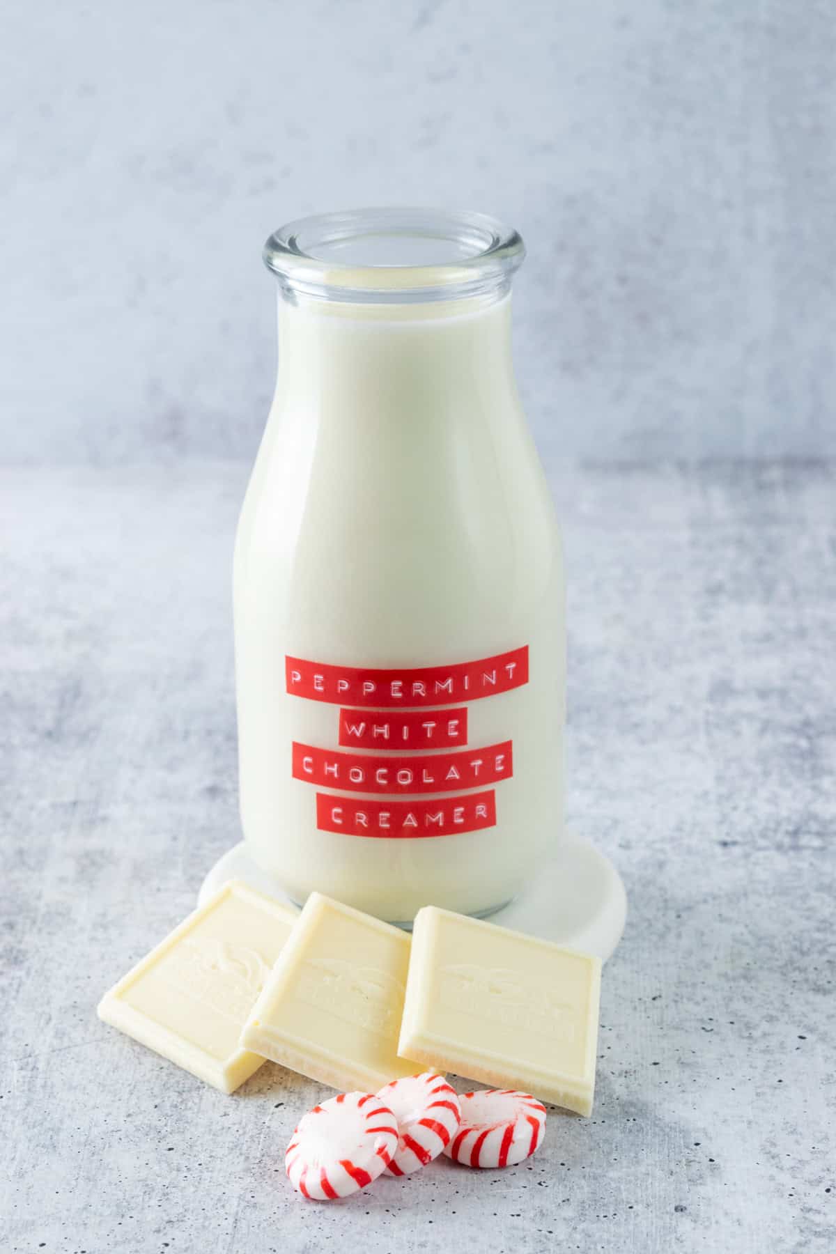 Bottle of homemade peppermint white chocolate coffee creamer with a red label on it, next two three squares of white chocolate and a few peppermint candies.