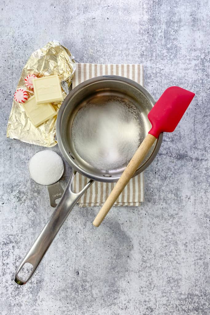 Sugar in a saucepan next to a few squares of white chocolate bar and peppermint candies.