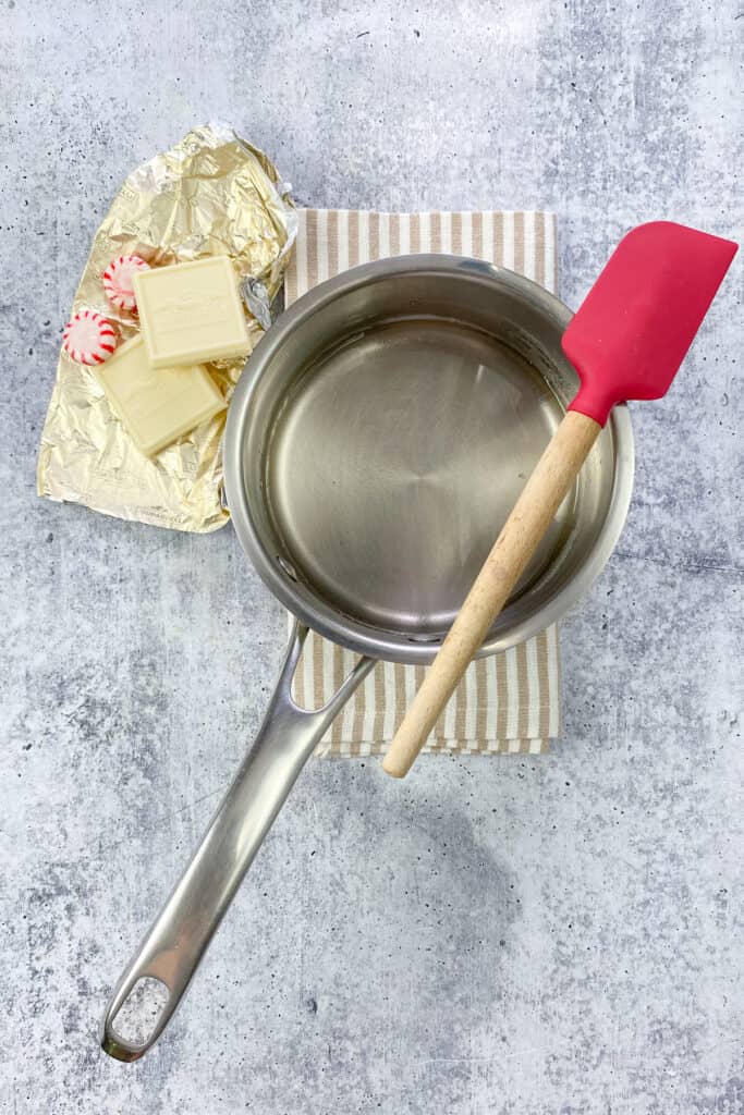 Saucepan containing clear simple syrup after sugar dissolves over heat, next to some white chocolate bar and starlight peppermint candies.