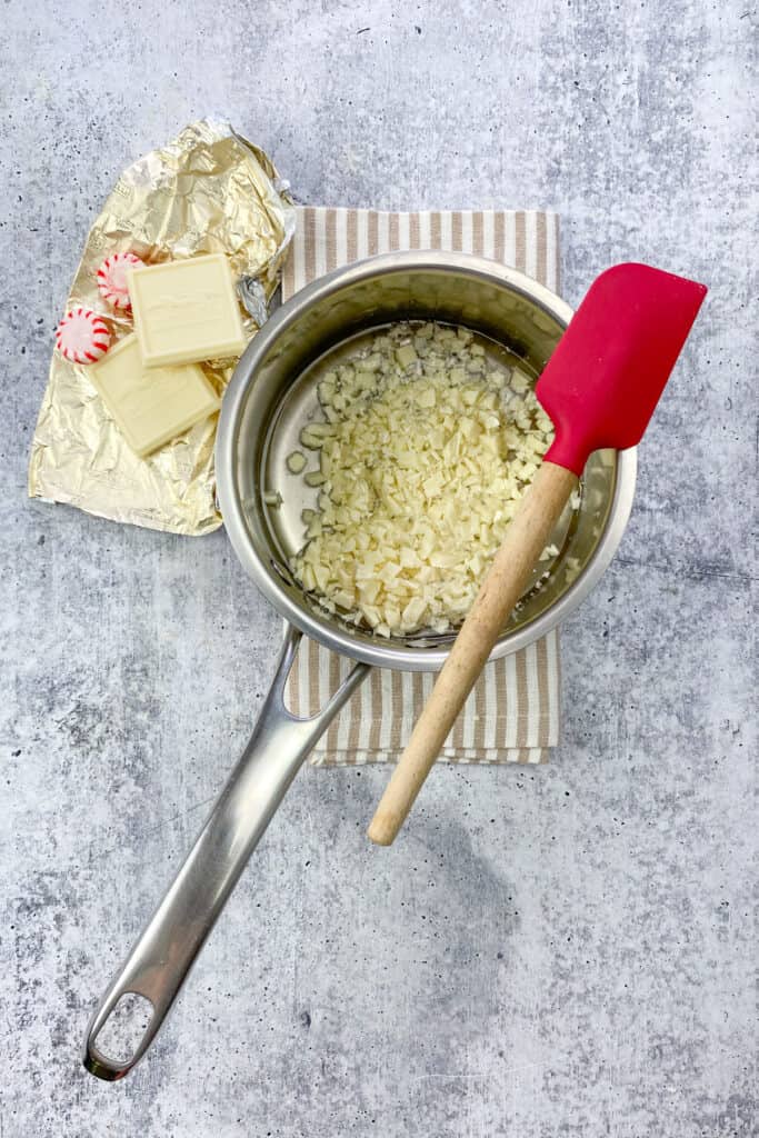 Evenly chopped pieces of white chocolate bar in the saucepan with the simple syrup with a red rubber spatula resting on top of the saucepan.