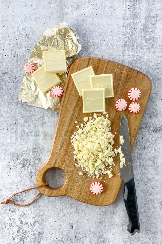 Ghirardelli white chocolate bar cut into small pieces on a wooden cutting board, next to a few squares of Ghirardelli white chocolate on the foil candy bar wrapper.