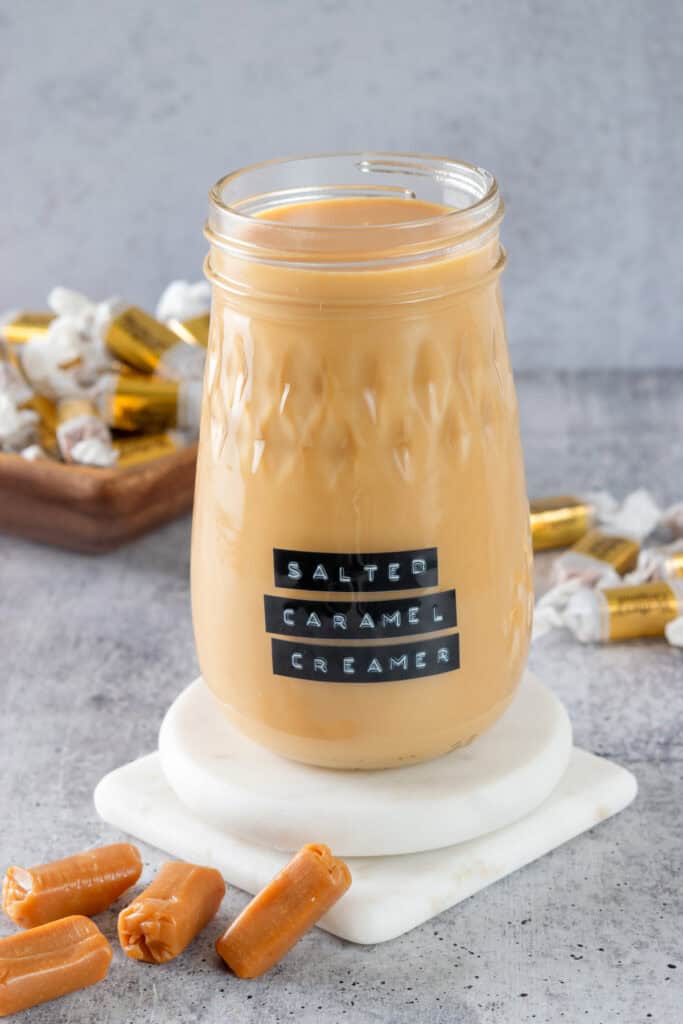 Bottle of homemade salted caramel coffee creamer with a label on it that says, salted caramel creamer. The jar is on a white coaster with caramel candies next to it.