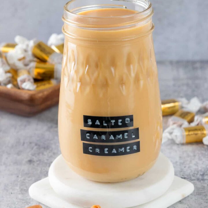 Bottle of homemade salted caramel coffee creamer with a label on it that says, salted caramel creamer. The jar is on a white coaster with caramel candies next to it.