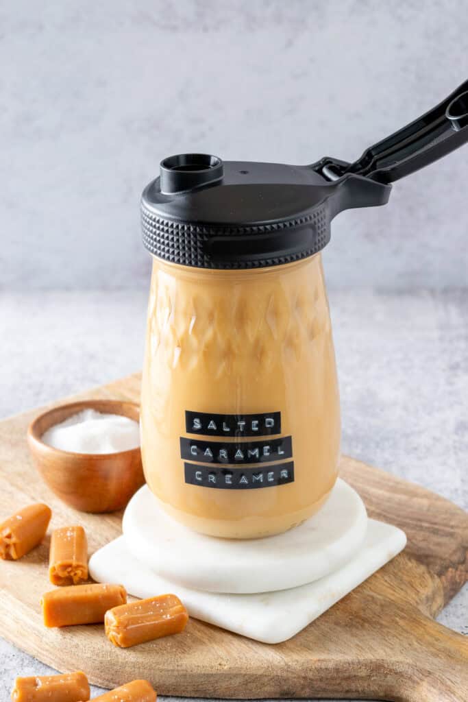 Glass jar full of homemade salted caramel creamer, sitting on a white coaster which is on a wood cutting board next to a small bowl of salt and a few caramel candies.