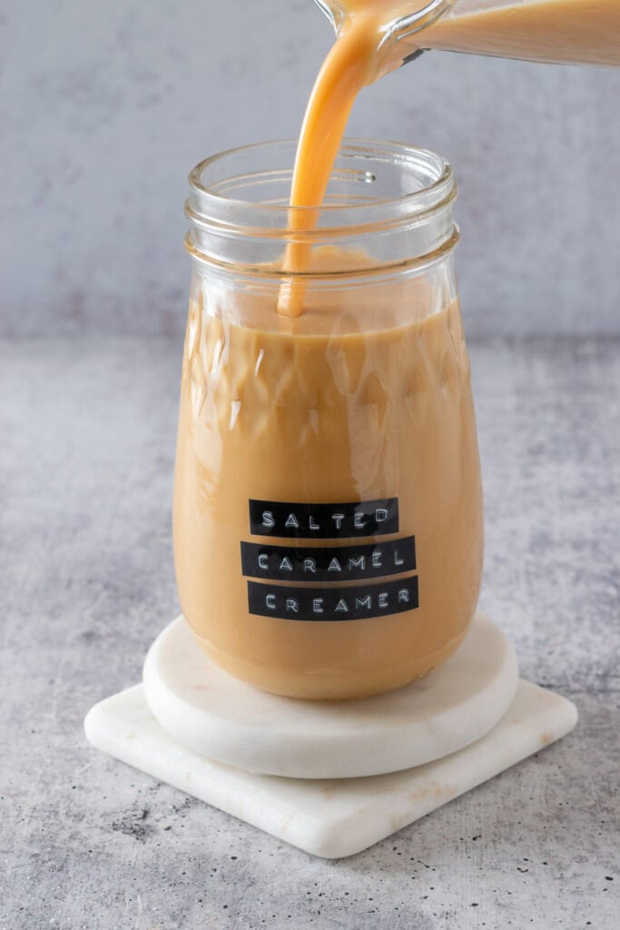 Pouring homemade salted caramel creamer from the measuring cup into a glass jar that has a label on it with the text, salted caramel creamer.