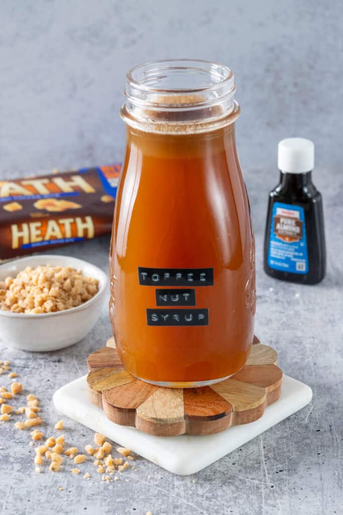 A bag of Heath toffee chips, bottle of pure almond extract, and small bowl with toffee bits in it, all next to a bottle of homemade toffee nut syrup.