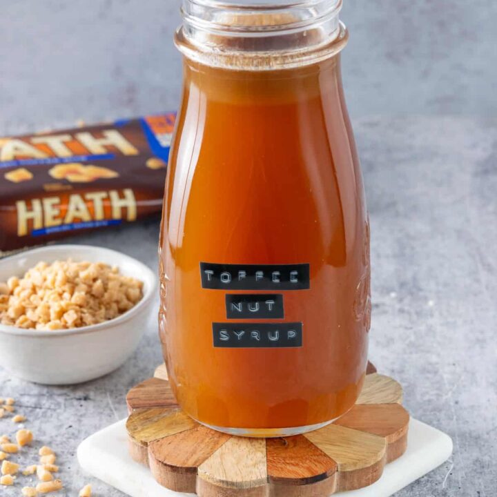 Bottle of homemade toffee nut syrup with a label on it, next to a bag of Heath toffee chips and a small bowl with toffee in it.