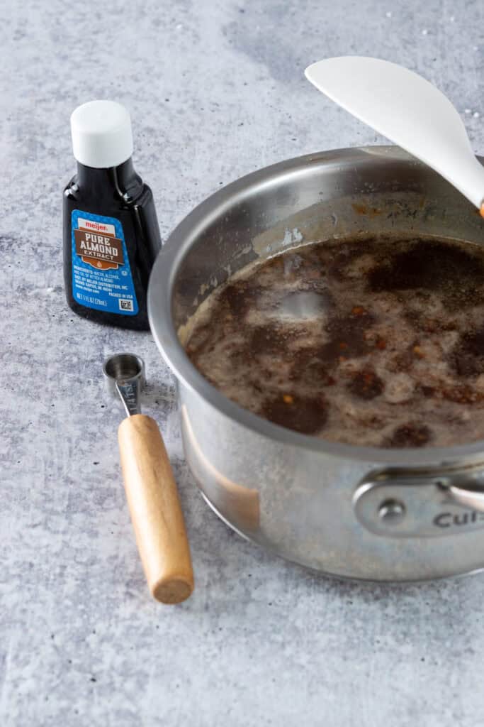 Homemade toffee nut syrup in a saucepan next to a bottle of pure almond extract and a 1/4 teaspoon measuring spoon.