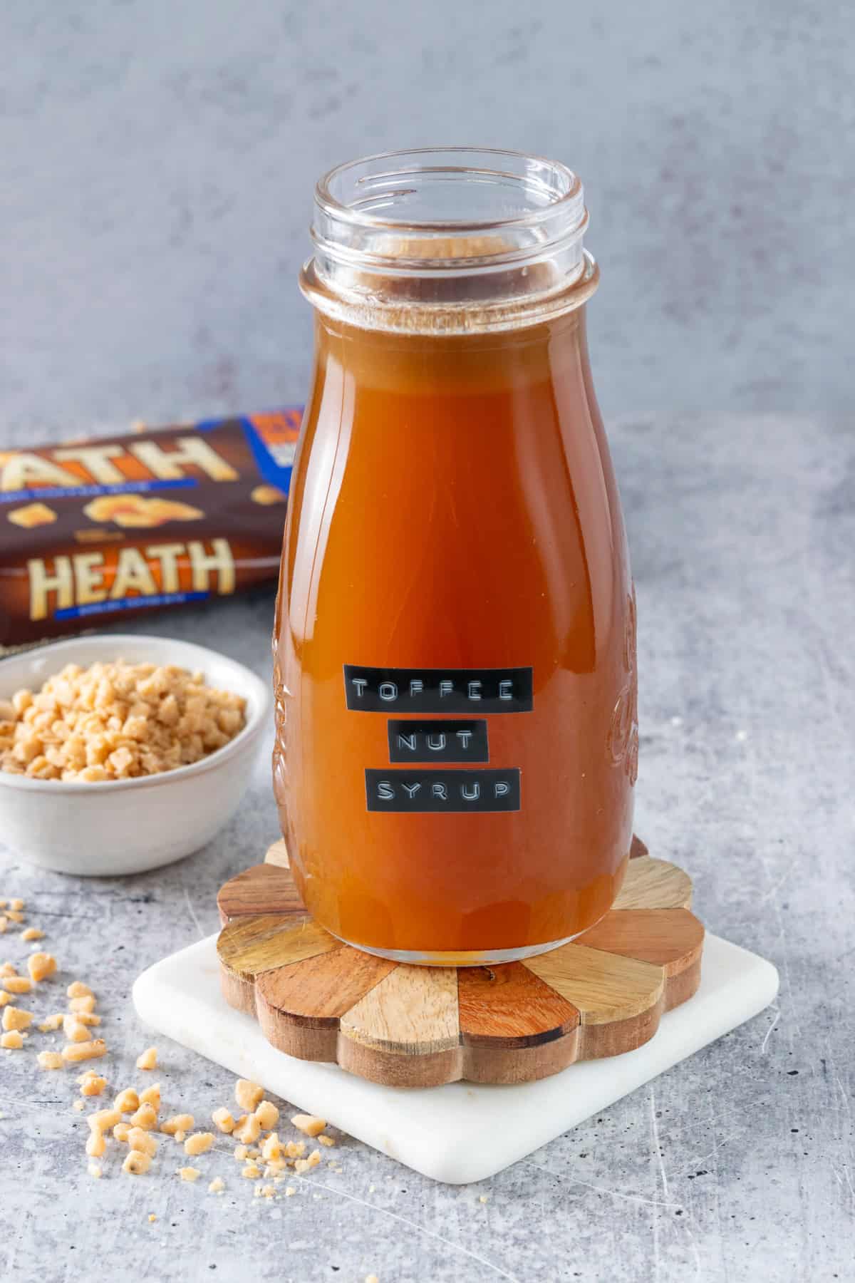 Bottle of homemade toffee nut syrup with a label on it, next to a bag of Heath toffee chips and a small bowl with toffee in it.