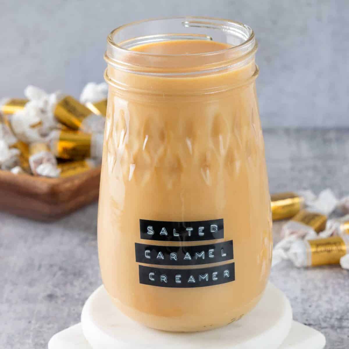 Bottle of homemade salted caramel coffee creamer with a label on it that says, salted caramel creamer. The jar is on a white coaster with caramel candies next to it.